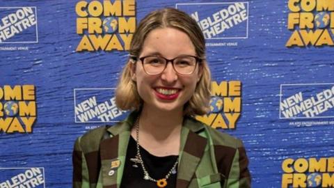 Lydia standing in front of Come From Away branding at the New Wimbledon Theatre. She has blonde hair, is wearing glasses and has red lipstick on. She is wearing a green checked jacket, a black t-shirt underneath and a necklace.