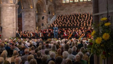 A choir stands behind an orchestra in a cathedral. The audience can be seen from the back. A man in a blue suit holds a microphone. Stained glass windows and pillars are visible in the background.