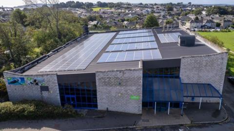 A drone shot of Saltash Leisure Centre. It is a square grey building and its roof is covered in solar panels. The entrance is glass with clear plastic panels above it. There are two posters marketing the facilities on one of the exterior walls. In the background there are residential houses and there is a flat green area of grass to the right of the centre.
