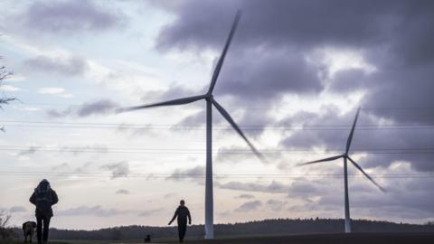 Two tall, white wind turbines are spinning in the wind. They are in a field with a line of trees in the distance. Two people in the foreground are walking their dogs.