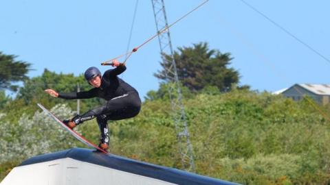 A person wakeboarding at a cable park. They're performing a trick on a white ramp in the water while holding onto a handle that's connected to an overhead cable system. In the background, you can see green trees, and bushes.