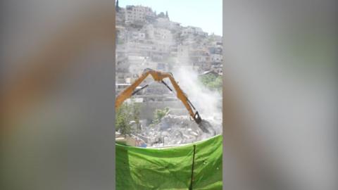 A yellow bulldozer engaging in the demolition of homes. It is behind a raised green net and behind it are other houses.