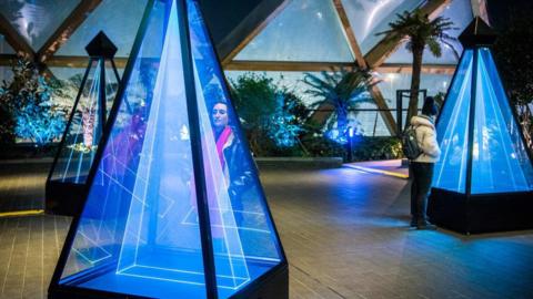 CGI images of three blue isosceles pyramids. They are made of glass and filled with blue lights. There are in an empty room. Two people are looking into two of them. There are triangular windows on the back wall which show the sky through them. The sky is blue 