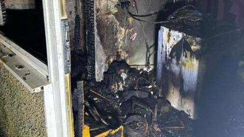 The burnt out remains of the washing machine with blackened wiring and soot covering everything.