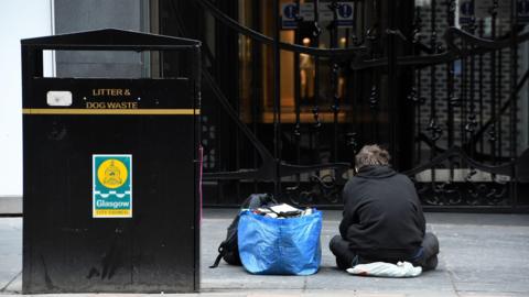 A homeless man wearing dark clothes sits next to a blue bag with his belongings and a large back bit on a city street.
