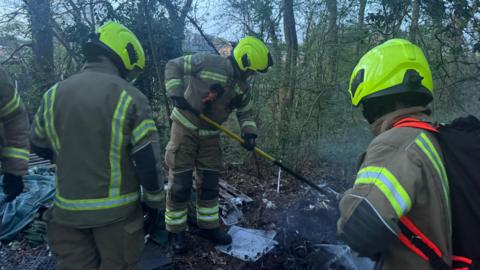 Fire crews in protective equipment look at the embers of a fire in woodland. 