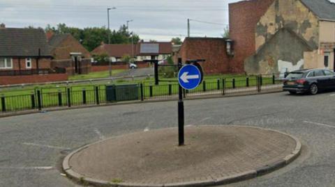 A roundabout connecting High Street and Elemore Lane in Sunderland. A black car is travelling away from the camera. A number of brick buildings can be seen to the side of the road.
