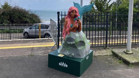 The statue has pink hair and a blue tail and is combing her hair. She is sitting on a green GWR pedestal. There is railway fencing behind the statue and a car park with one car behind that. On the horizon, there is a the beach looking over to a lighthouse.