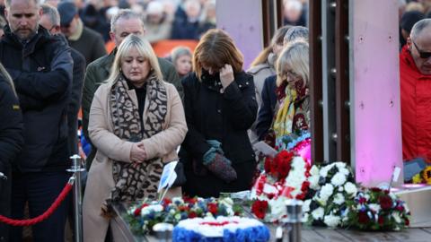 Several people gather around a memorial where multiple wreaths of flowers have been laid, in the centre of the image a woman is seen crying