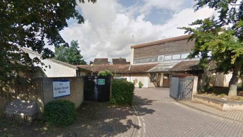 A brick and wooden-clad two-storey building standing inside a gated driveway. There is a sign on the wall saying "Ecton Brook House"