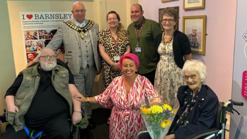 A photo of Peter and Patricia at the exhibition. They're joined by the Mayor of Barnsley, Councillor David Leech, Mayoress Alison Leech, Deansgate Care Home's events coordinator Rachael Addy, local artist Charlotte Fairclough and Barnsley Museums community heritage curator Steven Skelley.