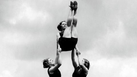 A black and white image of two women holding a third woman up in the air while an acrobatic exercise. They are wearing black leotards. The woman at the top of the image has her legs up in the air. 