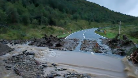 A mudslide has blocked a road with debris and flowing water, surrounded by lush greenery and a winding road visible in the background.