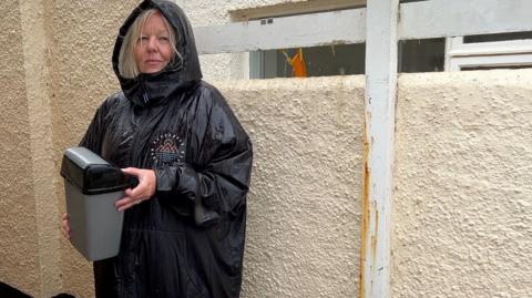 A woman in a raincoat carrying a small bin next to a line of large bins outside a public toilet.