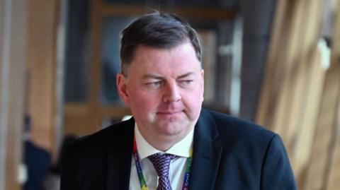 Colin Smyth, a man with flat brown hair, while wearing a suit, white shirt and purple tie and multicoloured lanyard
