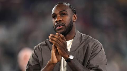A man standing on a pitch with a blurred purple light in the background. He has black hair in cornrows and a closely trimmed moustache and beard. He is smiling and looking into the distance. He is wearing a brown bomber jacket, cream t shirt and numerous gold and silver necklaces with charms.