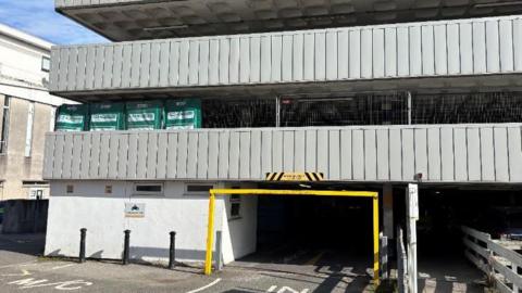 The ground level entrance to the car park has a yellow height restriction barrier and road markings saying In and M C, standing for motor cycles. Two more storeys can be seen above withy grey walls.
