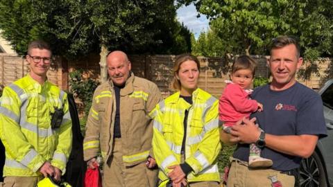 Four firefighters standing in a line, with the last one holding a girl in a pink top and floral leggings who looks to be around 18 months old