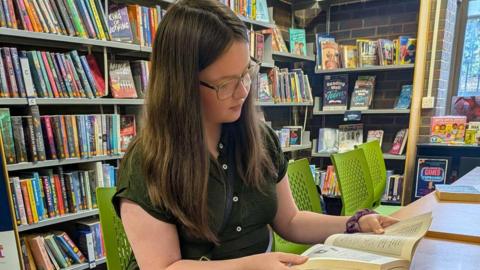 12-year-old Kaitlin reads a book in a library, sat at a desk on a bright green chair. She has long brown hair and wears a green short sleeve shirtdress. Behind her, shelves of brightly coloured library books can be seen.