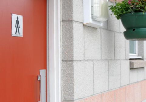 Image of the red door of a gents' public toilet, with a light and a hanging basket on the wall nearby
