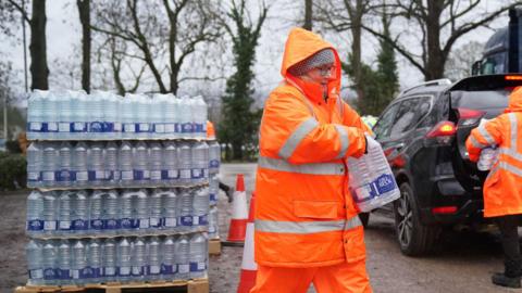 A woman in an orange jacket and trousers lift water bottles at a water station