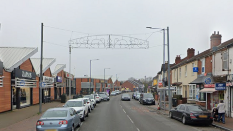The street has cars parked on either side with shops and houses also on both sides of the street. A Christmas decoration of lights can be seen hanging above the road.