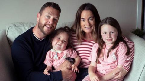 A family of four sat on a sofa. Parents Greg and Vic are sat side by side with their two daughters on their laps. Greg has short dark hair and facial hair and is wearing a black jumper. Vic has long dark hair and is wearing a red and white striped t-shirt. Margot is a four-year-old girl and she is sat on Greg's lap. She has dark hair and is wearing a pink t-shirt. Mimi is a six-year-old girl sat on Vic's lap. She also has long dark hair and a pink t-shirt on. All of them are looking at the camera and smiling. 