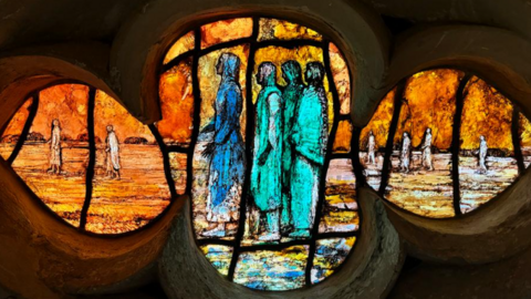 A close-up of a pane of the glass in a flower shape with several biblical figures depicted in hues of orange, turquoise and blue