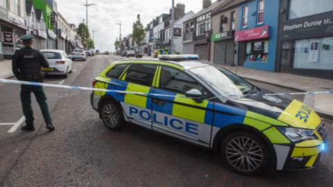 A police officer stands with their back to the camera beside a marked police car. There is a cordon in place sealing off a large main street road. There are a number of shops lining either side of the road, some with their shutters down.