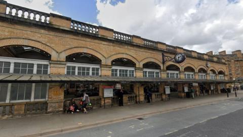 A railway station building with a clock hanging outside the front. People are sat on benches outside the building. A bus stop is marked on the road outside the building.