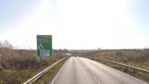 The A45 in Northamptonshire. It is single-carriageway and there is a green sign on the left which shows markings for an up-coming roundabout with directions to Northampton, Upper Heyford and Flore