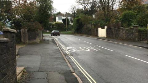Dean Hill in Plymstock, Plymouth. A black VW car is driving on the left-hand side of the road. Houses are either side of the road. The word "slow" is written in large white block capitals on the road, which has double yellow lines.