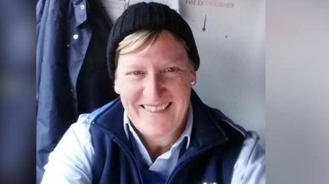 A woman with blonde hair poking out from under a black beanie hat grins. She is sitting down and wearing a blue shirt and navy bodywarmer.