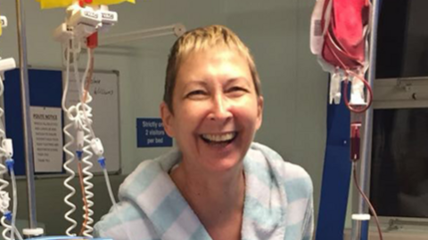 Paula Costin smiling in a light blue and white striped dressing gown standing in a hospital ward, flanked by various transfusion equipment