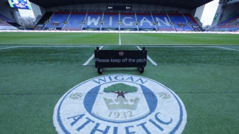 A view of Wigan's pitch with a Wigan Athletic badge on the artificial turf in front of the pitch and 'WIGAN' on the seats in the stand on the far side of the pitch 