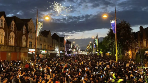 Crowds of people gather in the road as fireworks light up the sky
