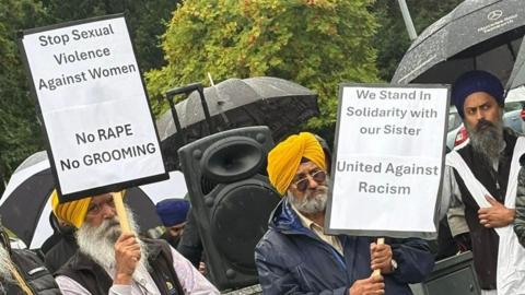 Two men wearing yellow dastars/turbans hold up large white signs that read "stop sexual violence against women. No rape. No grooming" and "We stand in solidarity with our sister. United against Racism".