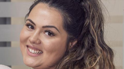 Close-up of Ella McKenzie smiling at the camera side-on. She has long dark hair in a ponytail.