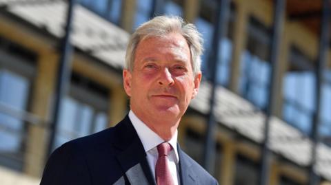 A man in a suit and tie, looking at the camera with a neutral expression. Rows of windows on an out-of-focus building can be seen behind him.