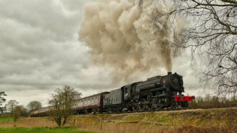 An old train on the Churnet Valley Railway emits steam as it travels along the track through the countryside.