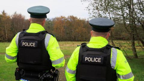 Two PSNI officers have their backs to the camera. They have on yellow police coats and black body armour that says 'police' on the back.