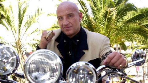 An image taken in Cannes in 2001 shows a younger Dave Courtney, a man with a bald head wearing a light jacket and black shirt, sitting on a motorbike with a close-up view of three round silver headlights on the handlebars. Small palm trees can be seen in the background