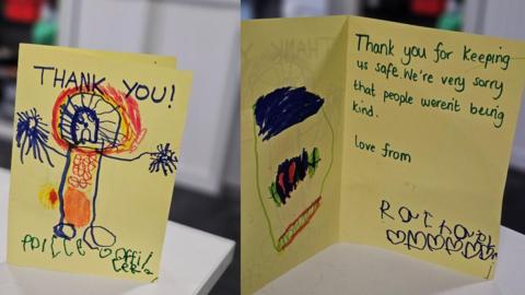 Two cards made on yellow paper by a young child with a child's picture of a police officer and the words "thank you" on one and the words "thank you for keeping us safe - we're sorry that people weren't being kind" on the other.