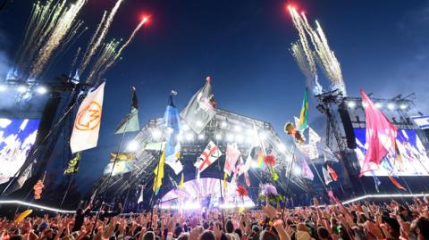 Thousands of fans are pictured in front of the Pyramid Stage at Glastonbury 2024. Many flags are visible and there are fireworks streaking into the sky