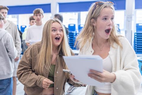Photograph of two GCSE students receiving their exam results. One girl (right) holds her papers in her hand and has her mouth open in shock. She has blonde hair and wears a cream cardigan and sunglasses on her head. Another girl stands beside her on her left. She has long blonde straight hair and also looks shocked. She wears a brown trucker jacket.