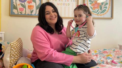 A woman with dark hair, wearing a bright pink jumper holds her three-year-old daughter on her lap. She is wearing a striped t-shirt with a pink toy stethoscope held up to her ears.
