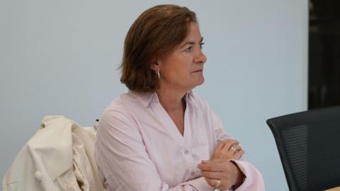 Eluned Morgan sat at a table on a chair with a binder in front of her, and a jug of water. She has her arms crossed and has a neutral expression.