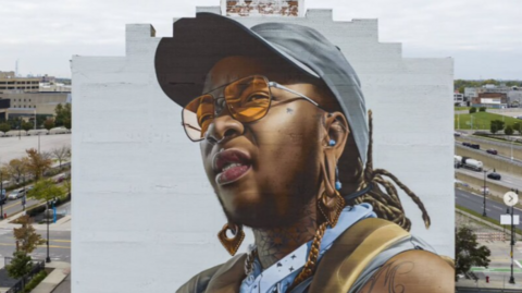 A mural has been painted on the side of a building. It is of a person wearing a grey baseball cap and orange sunglasses.