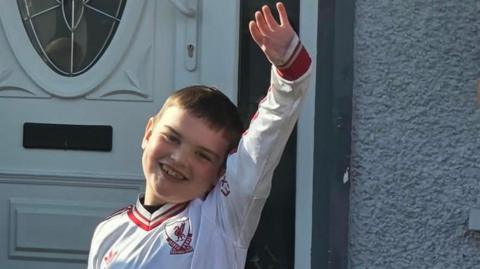 Dáithí Mac Gabhann. He is smiling and holding his hand up in the air. He is wearing a white long sleeve Liverpool jersey. A white door is behind him.