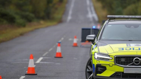 A police car is parked within a coned-off section of a single carriageway road.